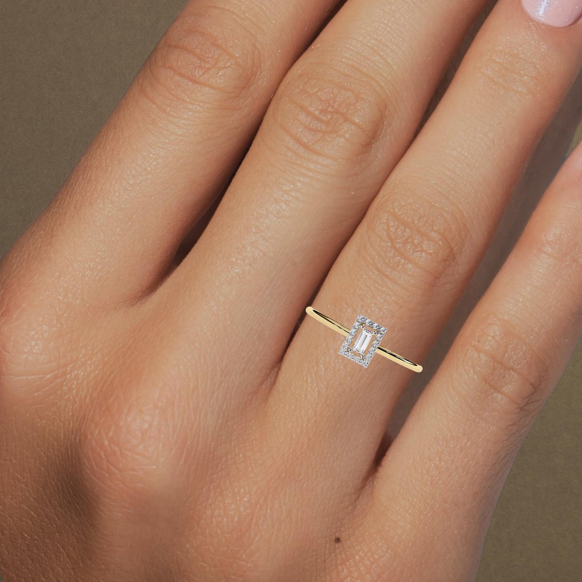 A yellow gold ring worn on a finger, featuring a rectangular baguette-cut diamond at the center, surrounded by a halo of round brilliant diamonds. The diamonds are set in a sleek, minimalistic band, highlighting the elegance of the baguette center stone. The simple yet sophisticated design adds a modern touch to this timeless piece, making it a perfect engagement ring or a refined statement piece.