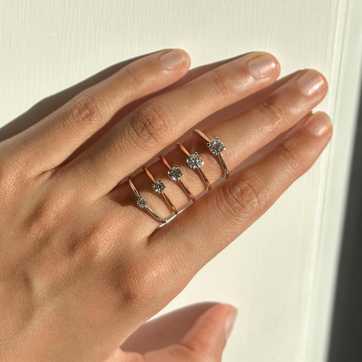 Hand wearing five minimalist solitaire rings in 18K solid gold, each set with a single round lab-grown diamond in varying carat weights (0.10ct to 0.50ct). Dainty prong-set design, perfect for everyday wear, promise or engagement. Available in white, yellow, and rose gold.