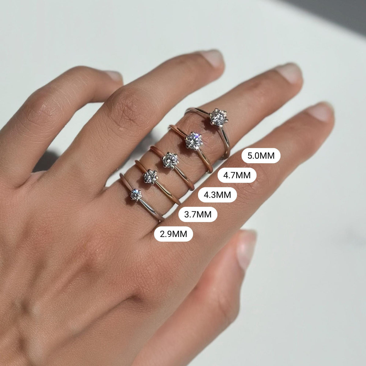 Hand showcasing a stack of six solitaire rings in 18K solid gold, each featuring a round cut lab-grown diamond in a classic six-prong setting; diamond sizes labeled from 2.9mm (0.10 ct) to 5.0mm (0.50 ct), offering a visual comparison of carat sizes—perfect as minimalist engagement rings, promise rings, or stackable fine jewelry gifts.