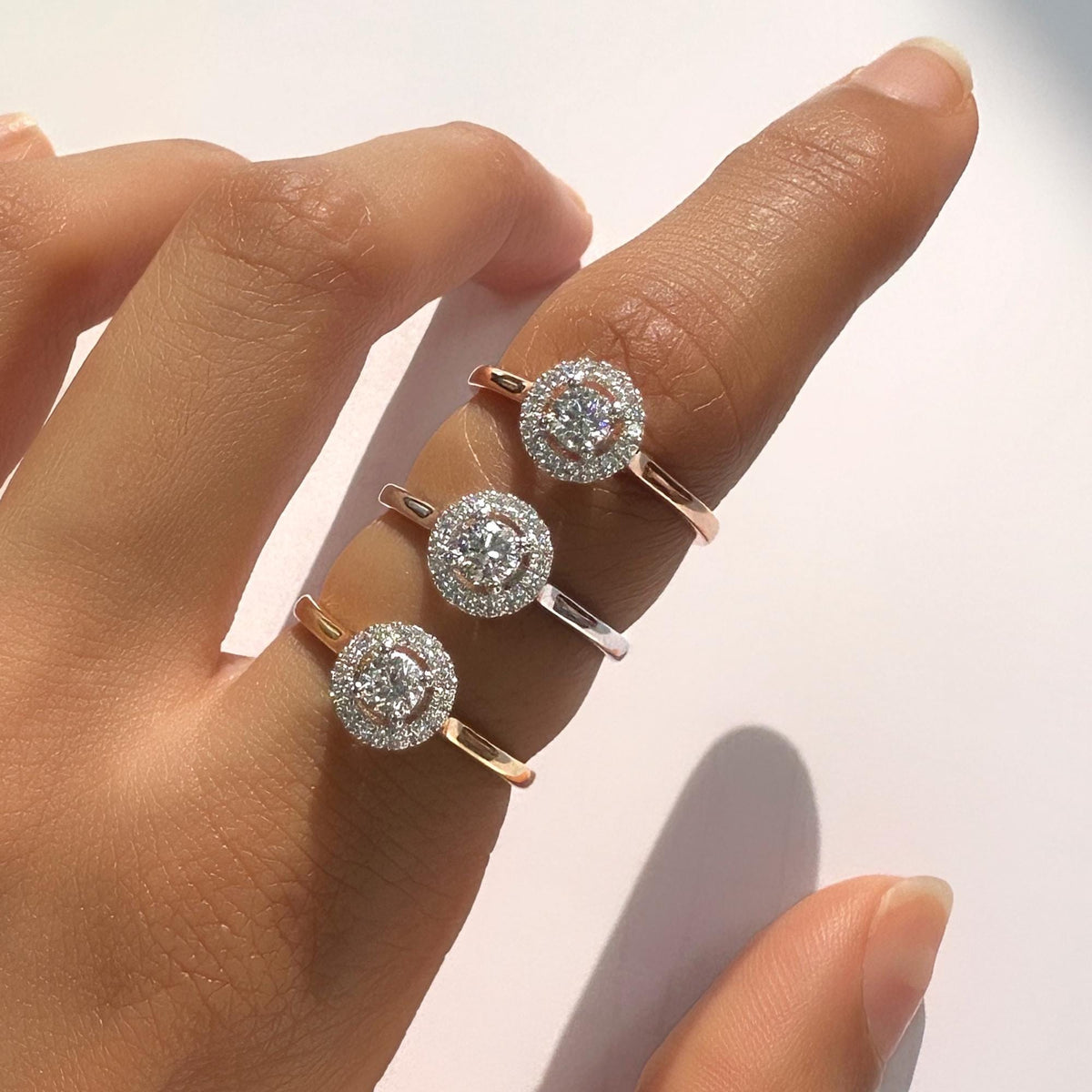 Three 18K Gold Halo Diamond Rings stacked on finger, each featuring a round lab-grown diamond in the center. Shown in yellow, white, and rose gold. Dainty minimalist promise and engagement ring set