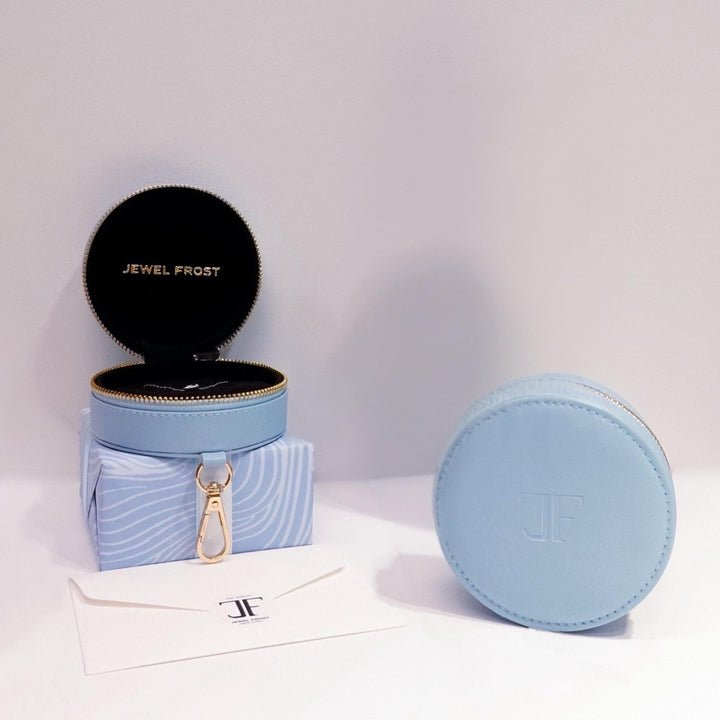 Jewel Frost light blue round travel jewelry case with gold zipper and clip, shown open and closed. The interior features a black velvet lining with JEWEL FROST embossed in gold. Displayed with a branded envelope and textured blue gift box for elegant presentation.