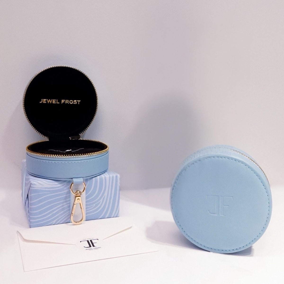 Jewel Frost signature packaging featuring a round light blue leather zip case with gold zipper, black velvet interior, and monogram "JF" branding. Includes a soft-touch envelope and gift-wrapped box, ideal for gifting fine jewelry.