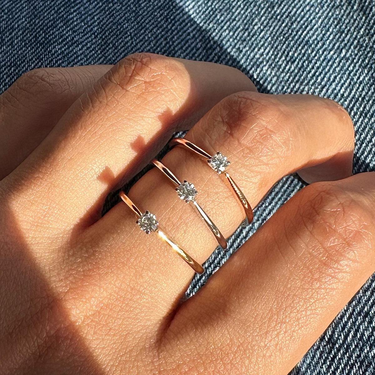 Hand wearing three 18k rose gold solitaire rings with round lab-grown diamonds in prong settings. Dainty 18K gold bands styled against denim for a minimalist everyday look.