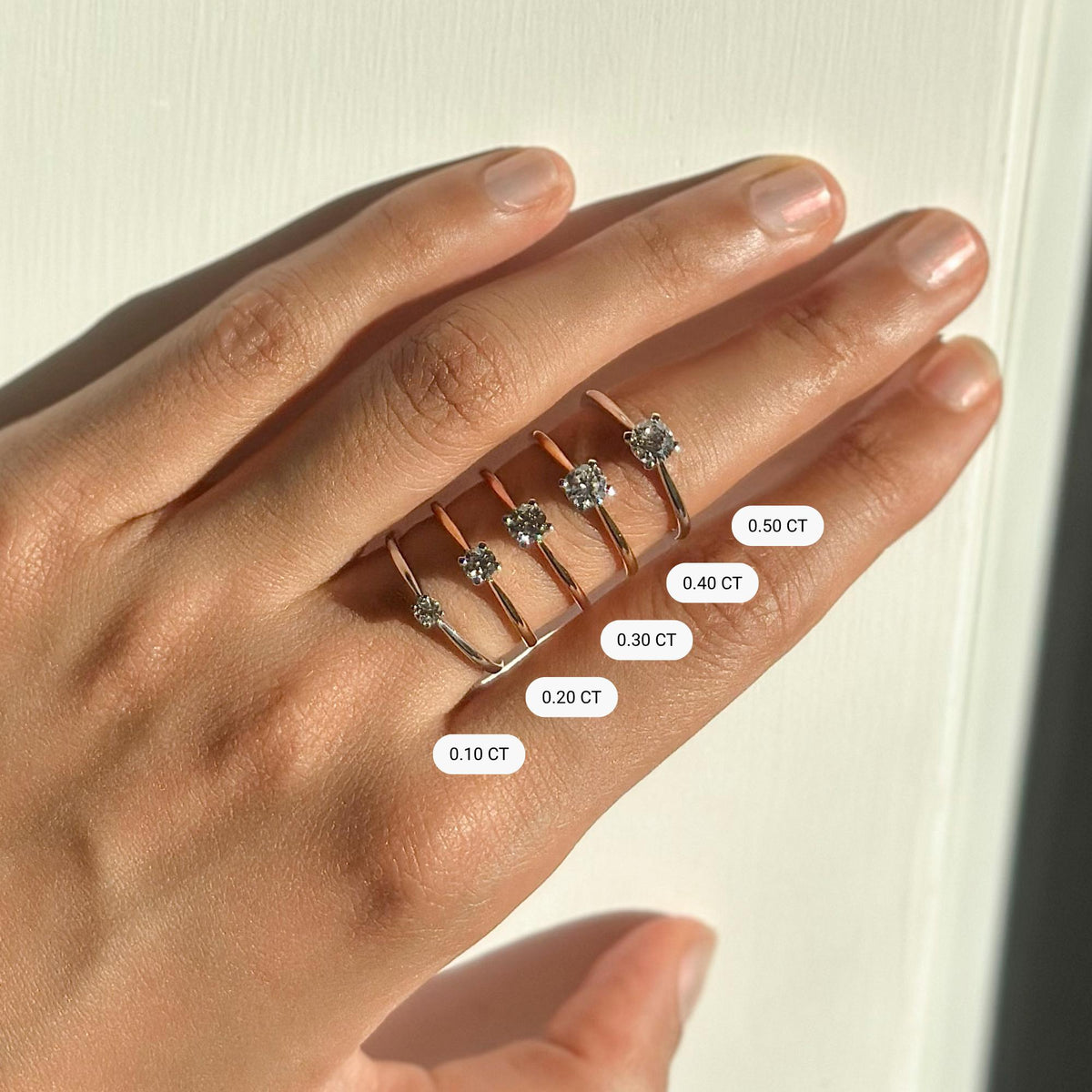 Five dainty solitaire diamond rings in rose and yellow gold displayed on a hand, labeled with carat weights from 0.10 to 0.50 CT. Visual comparison of minimalist diamond ring sizes for promise, engagement, or everyday wear.