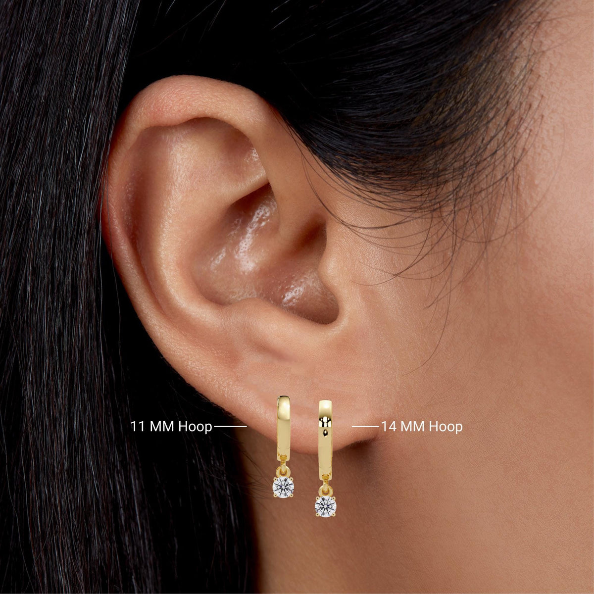 ear wearing two 14K yellow gold huggie hoop earrings with round lab-grown diamond drops in four-prong settings. Left hoop measures 11mm and right hoop measures 14mm, both shown for size comparison.