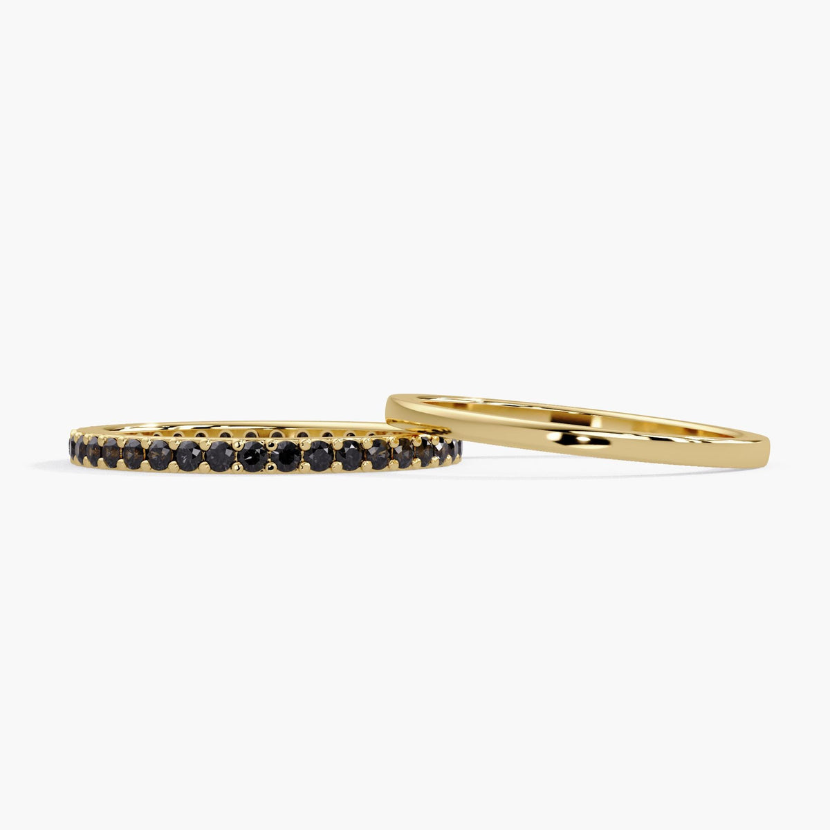 A side-by-side view of two 14K yellow gold rings: a stackable eternity band featuring 38 round-cut, natural black diamonds in a pave setting, next to a plain, thin, polished gold band.